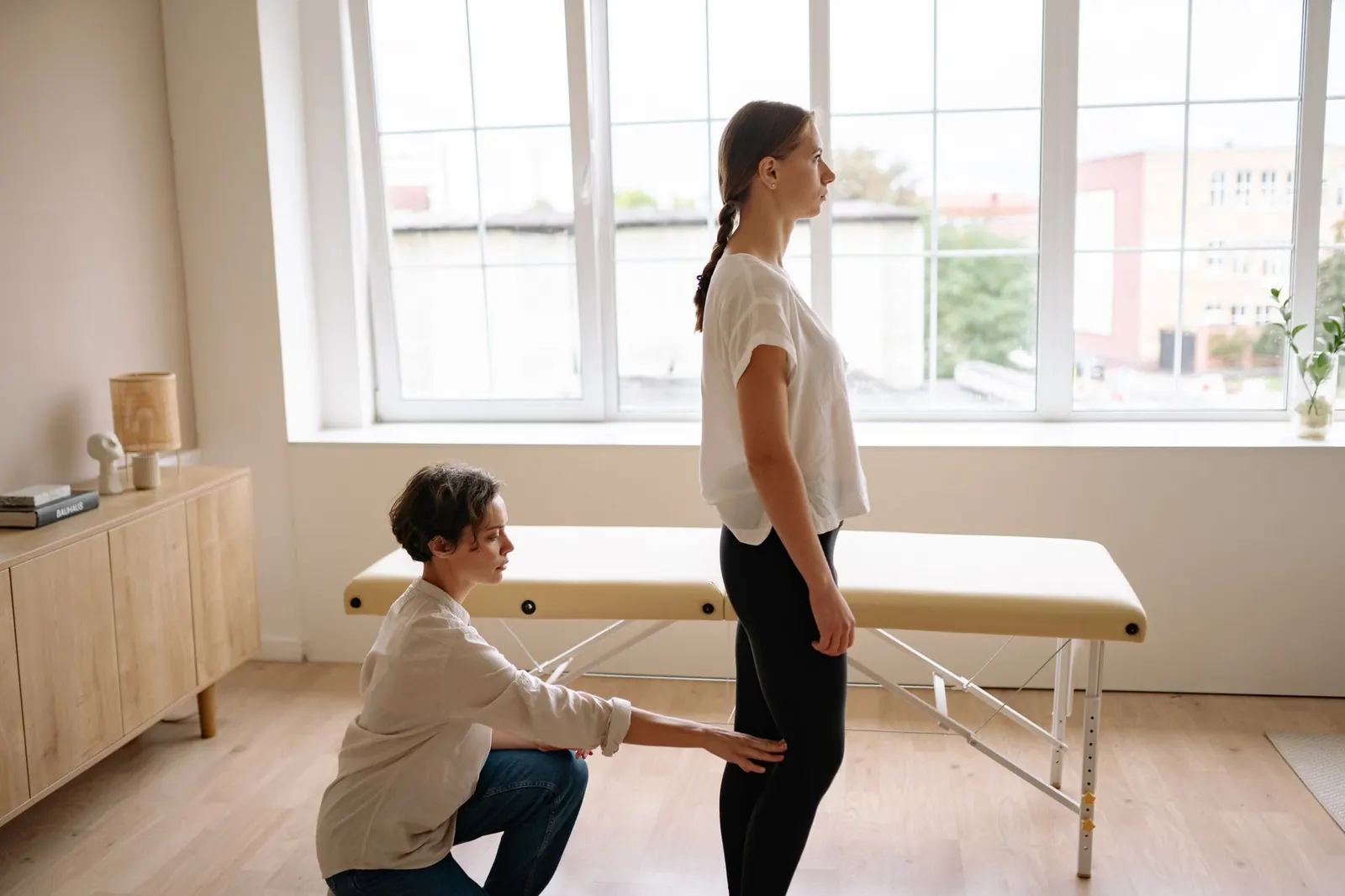 Calm, sunlit treatment room where a therapist assesses a standing patient